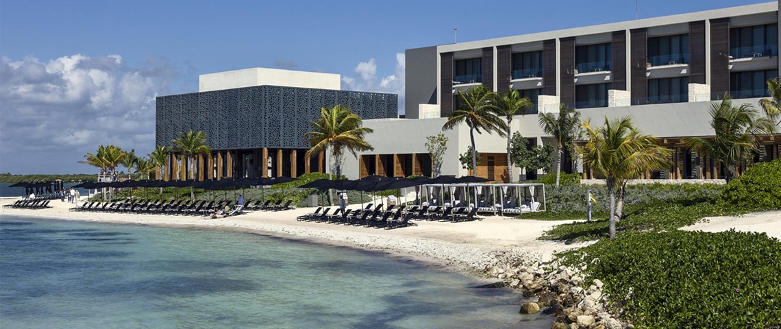 ビーチベッドとビーチチェアが並ぶ白い砂浜とモダンなホテル外観